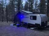 Night camping: A small grey RV rental with an extended awning and blue light, parked in a pine forest with coolers outside.
