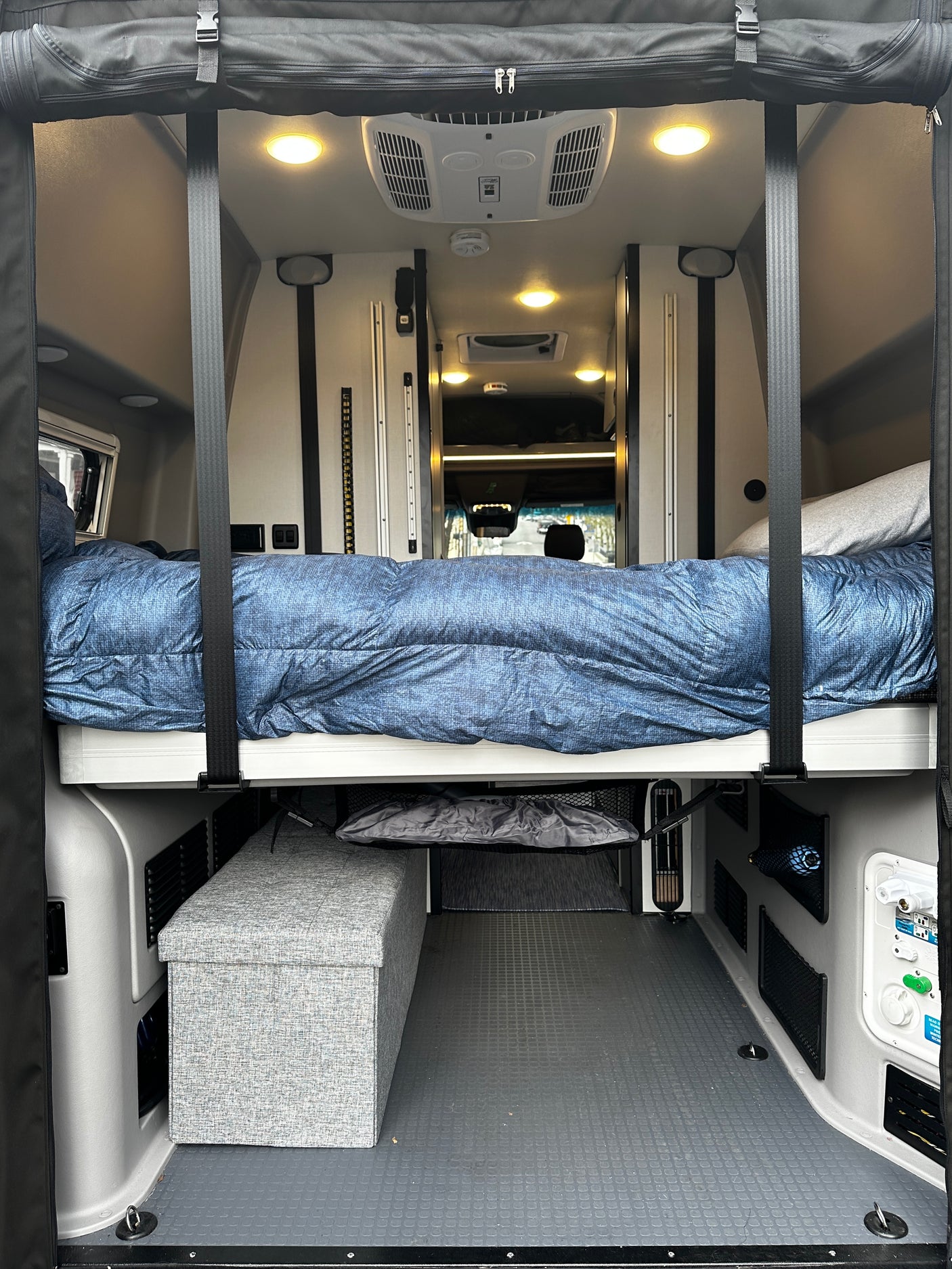 Step inside this RV rental! View of a comfortable bed suspended over storage & utility panels in a spacious camper van interior.