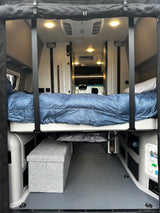 Step inside this RV rental! View of a comfortable bed suspended over storage & utility panels in a spacious camper van interior.