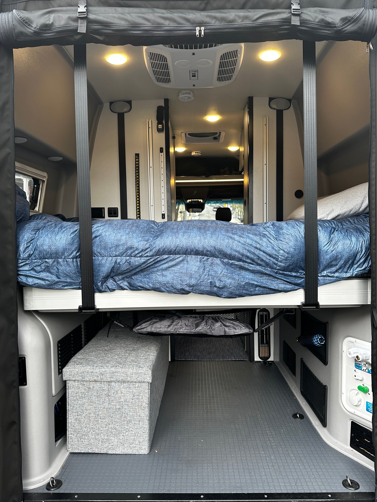 Step inside this RV rental! View of a comfortable bed suspended over storage & utility panels in a spacious camper van interior.