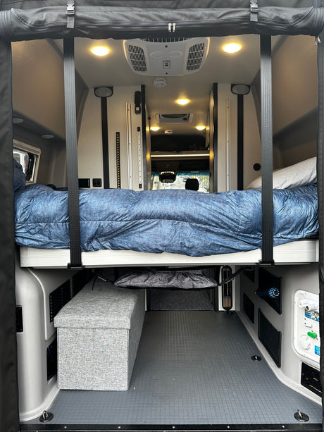 Step inside this RV rental! View of a comfortable bed suspended over storage & utility panels in a spacious camper van interior.