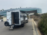 This white and black Wolf Pup travel trailer, an ideal RV rental for camping, has its door open and awning out in a parking lot.