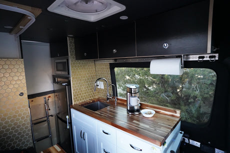 RV rental interior: Custom kitchen with wood counters, sink, microwave, and hex-tile backsplash. Trees visible outside.