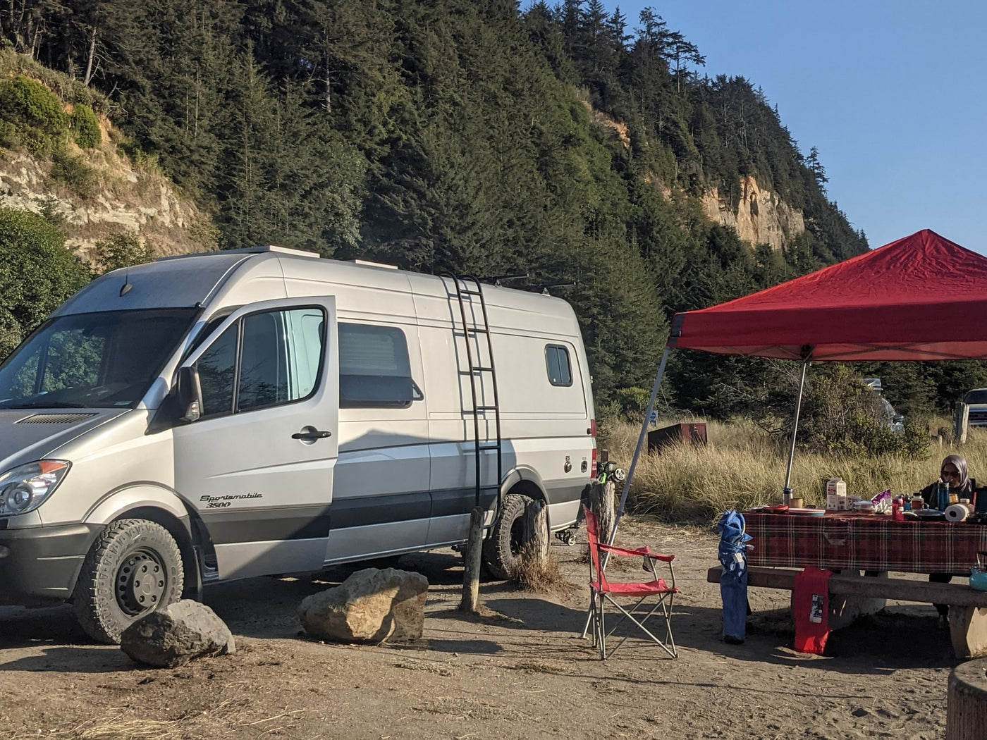  parked on a dirt path surrounded by tall pine trees in a sunny forest.|A silver Sprinter RV rental van on a dusty road — photo 4