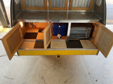  sits ready on a concrete lot near a larger RV and moving trucks.|Compact silver teardrop RV rental with yellow trim and fenders
