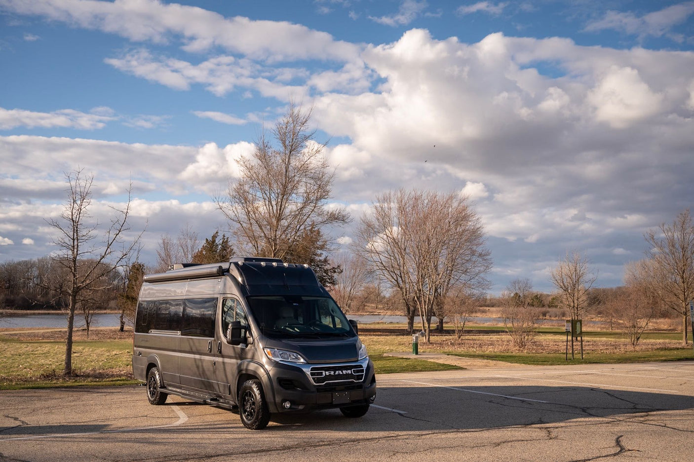 Dark grey RAM camper van, available for RV rental, parked by a lake with bare trees under a bright, cloudy sky.