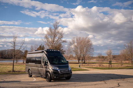 Dark grey RAM camper van, available for RV rental, parked by a lake with bare trees under a bright, cloudy sky.