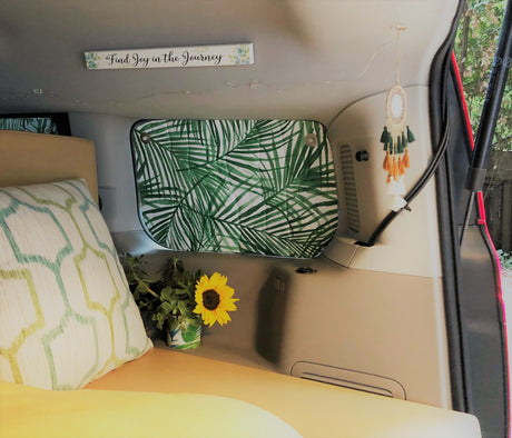 Cozy RV rental interior with a bed, palm leaf window cover, sunflower, dreamcatcher, and "Find Joy in the Journey" sign.