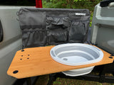 RV rental camping setup: A vehicle tailgate with a portable sink, wooden table, and fabric organizer with pockets.