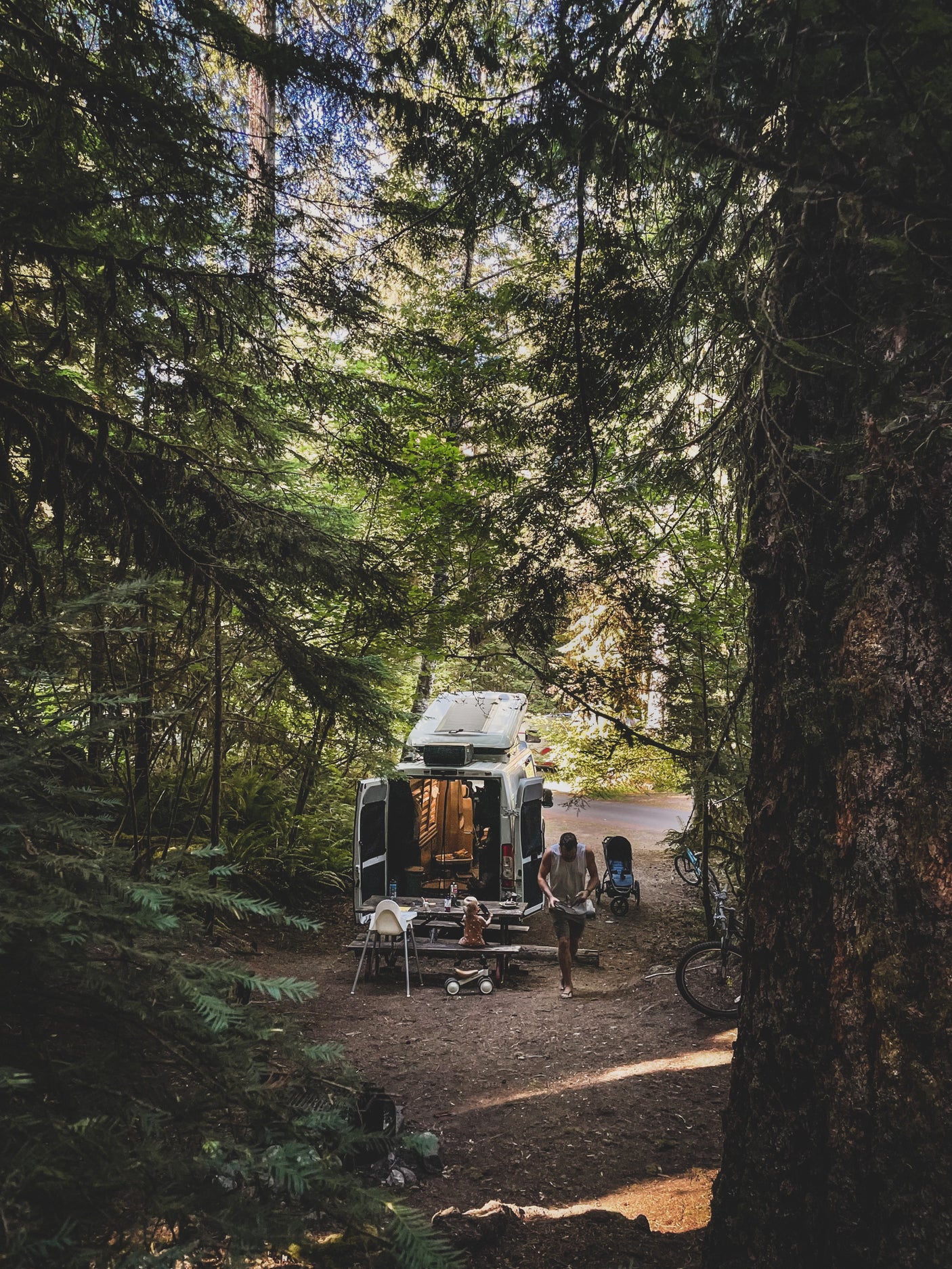 Forest campsite with an RV rental van, a man, and a child near a picnic table, enjoying the outdoors.