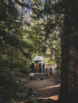 Forest campsite with an RV rental van, a man, and a child near a picnic table, enjoying the outdoors.