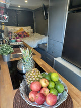 RV rental interior featuring a wooden counter with fresh fruit, pineapple, and a small plant, overlooking a cozy bed and storage.