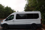 Side view of a white camper van, possibly an RV rental, with tinted windows and a roof solar panel, parked outdoors.