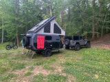 RV rental: Grey off-road camper trailer with rooftop tent & solar panels, hitched to a Jeep in a wooded campsite.
