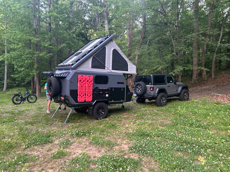 RV rental: Grey off-road camper trailer with rooftop tent & solar panels, hitched to a Jeep in a wooded campsite.