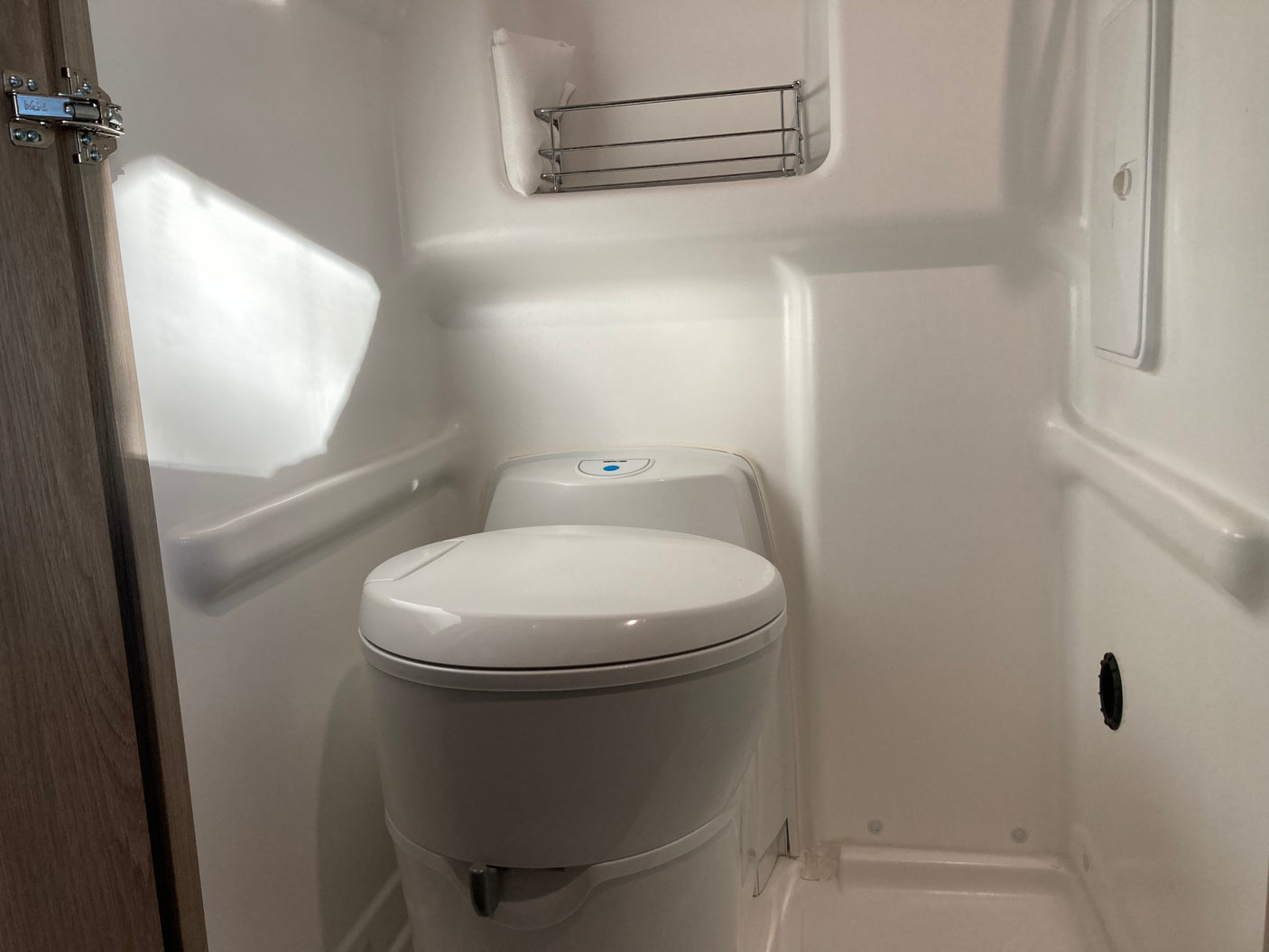 Compact RV rental bathroom with a white toilet (blue button visible) and a small wall-mounted metal shelf.