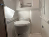 Compact RV rental bathroom with a white toilet (blue button visible) and a small wall-mounted metal shelf.