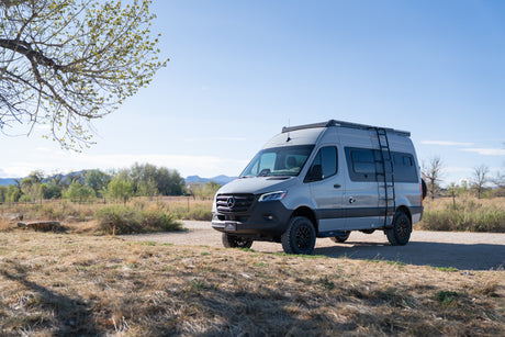 A grey Sprinter camper van, ideal for rv rental adventures, parked on a dirt path with a tree and mountains in background.