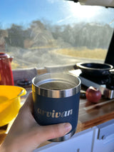 Cozy RV rental morning: hand holds steaming coffee inside, with kitchen setup & scenic view through the window.