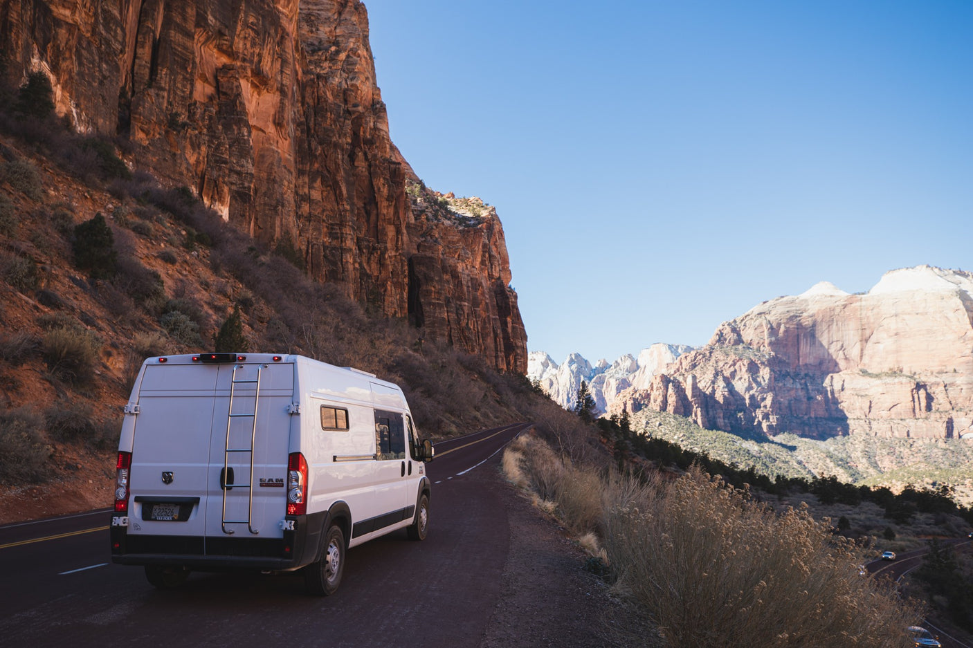 This RV rental van is parked on a winding road amidst the towering red cliffs of Zion National Park. Clear blue sky.