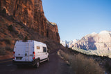 This RV rental van is parked on a winding road amidst the towering red cliffs of Zion National Park. Clear blue sky.