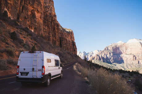 This RV rental van is parked on a winding road amidst the towering red cliffs of Zion National Park. Clear blue sky.