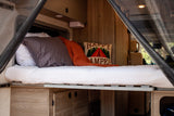 Cozy RV rental bed with "Happy Camper" pillow, showcasing comfortable interior sleeping and storage space.