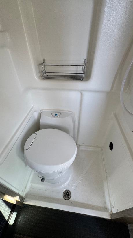Compact RV rental bathroom features a white toilet, integrated shower pan with drain, and wall-mounted shelf.