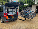 This RV rental van, tailgate open, shows organized storage bins & a yoga mat. Four mountain bikes on a hitch rack.