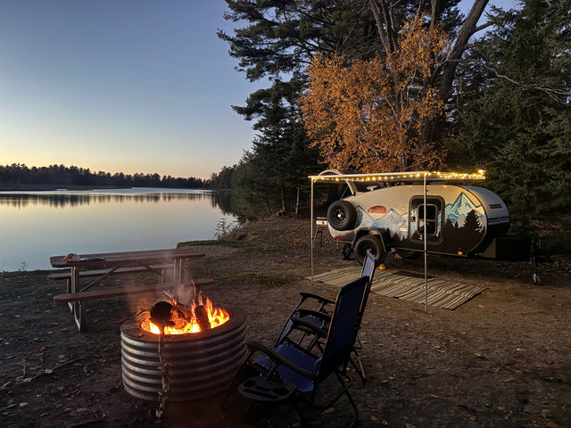 Enjoy a serene RV rental camping experience! A cozy campfire burns by a teardrop trailer and a calm lake at twilight.