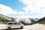 White camper van (RV rental) on a mountain road, with snow-covered peaks and evergreen forests under a blue sky.