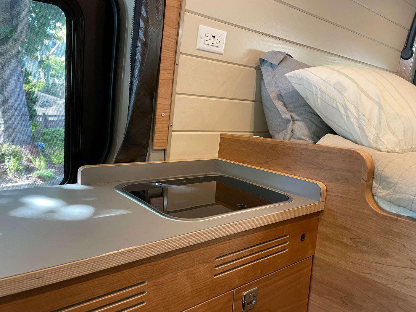 Explore this RV rental's interior: a cozy kitchen area with an integrated sink, wooden cabinets, and a comfortable bed by the window.