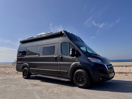 Dark gray RV rental camper van with all-terrain tires, parked on a paved lot by a sunny beach with the ocean and a ship in view.