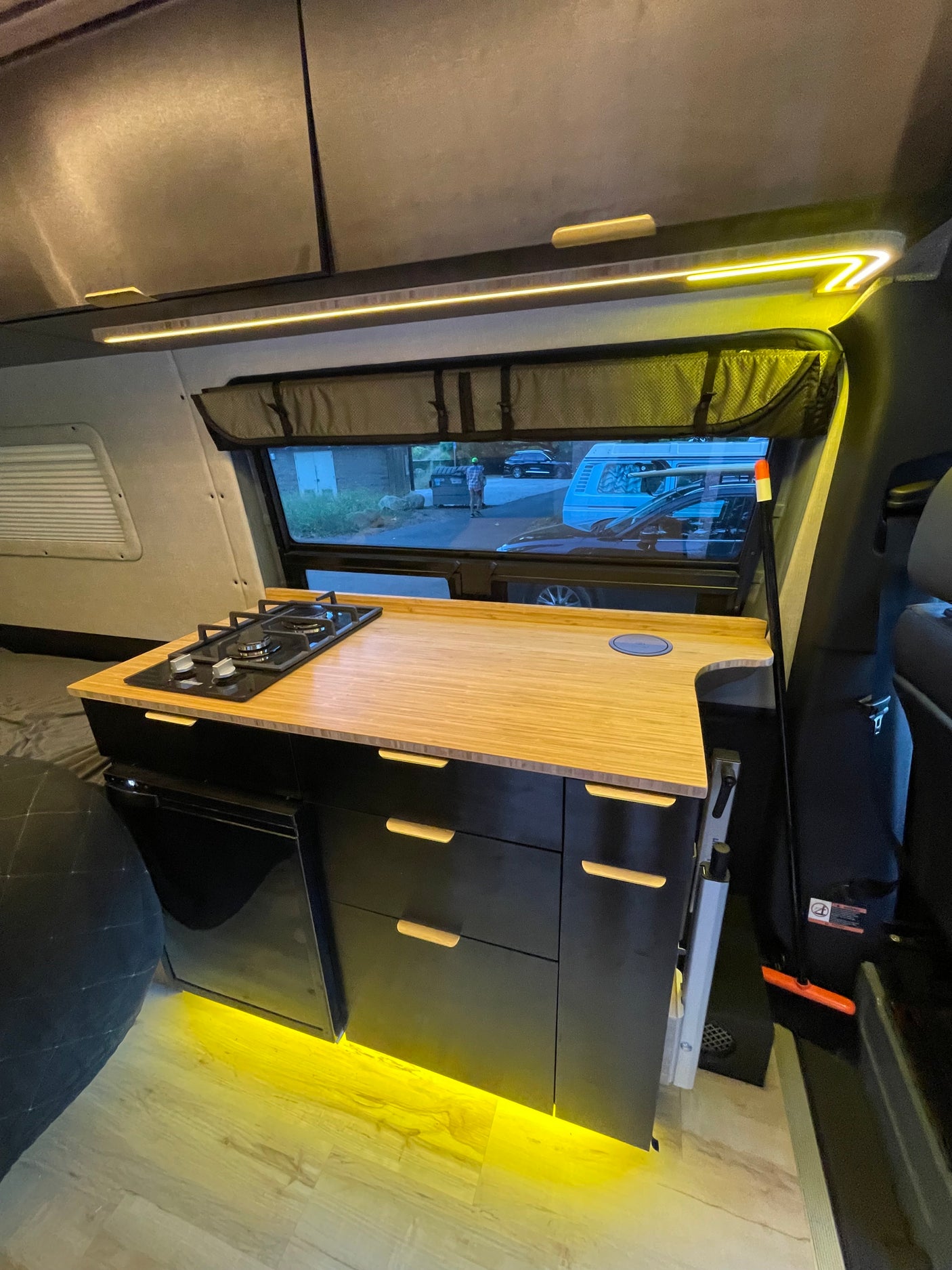 Compact RV rental kitchen featuring a gas stove, bamboo counter, fridge, & warm lighting. Perfect for mobile cooking.