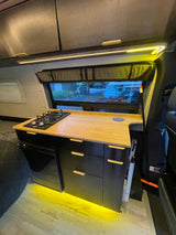 Compact RV rental kitchen featuring a gas stove, bamboo counter, fridge, & warm lighting. Perfect for mobile cooking.