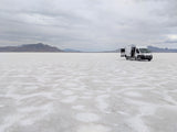 White RV rental van on vast Bonneville Salt Flats with open doors, mountains under cloudy sky.