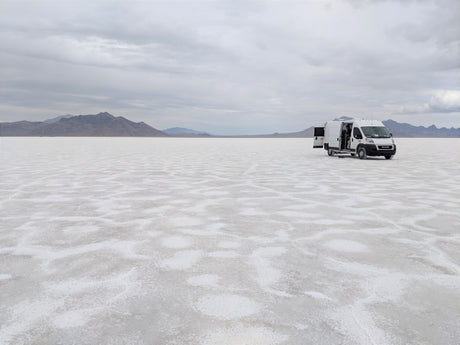 White RV rental van on vast Bonneville Salt Flats with open doors, mountains under cloudy sky.