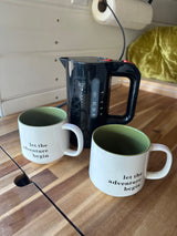 Start your day with coffee in your RV rental! Black kettle & 'let the adventure begin' mugs on a wood counter.