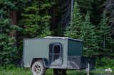 Mud-splattered green off-road RV rental trailer nestled in a dense forest, ready for adventure.