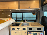 Compact RV rental interior featuring a bamboo kitchen, sink, cooktop, and cozy bed area, perfect for mobile adventures.