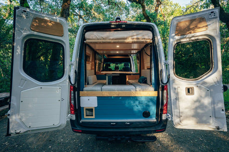 Open doors of an RV rental camper van showcase its cozy, converted interior with a bed and seating, parked in nature.