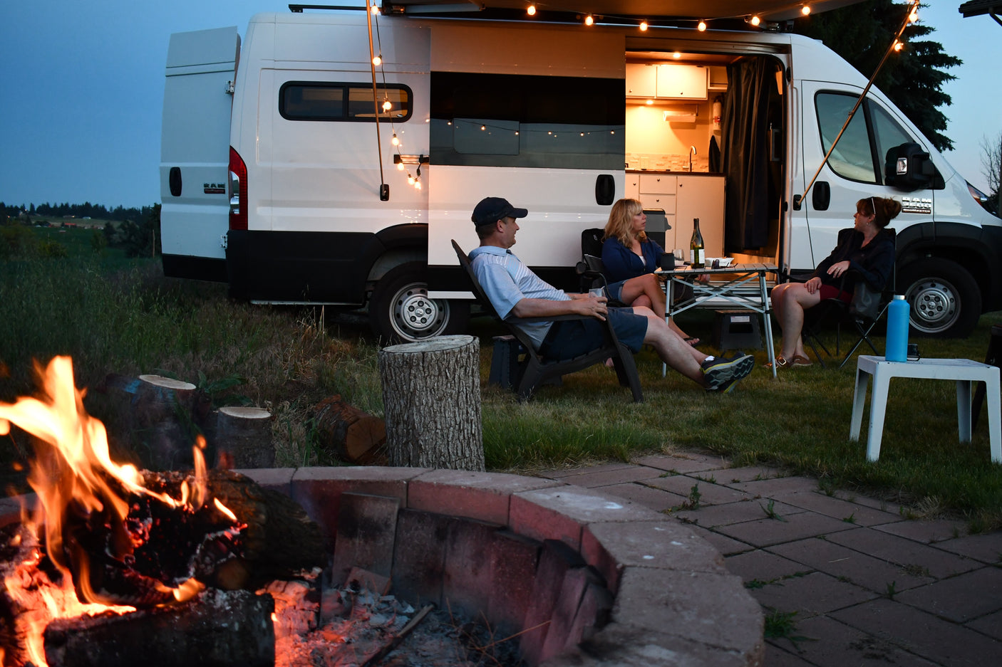 Friends relax by a warm campfire near their RV rental, showcasing illuminated van life on a peaceful evening.