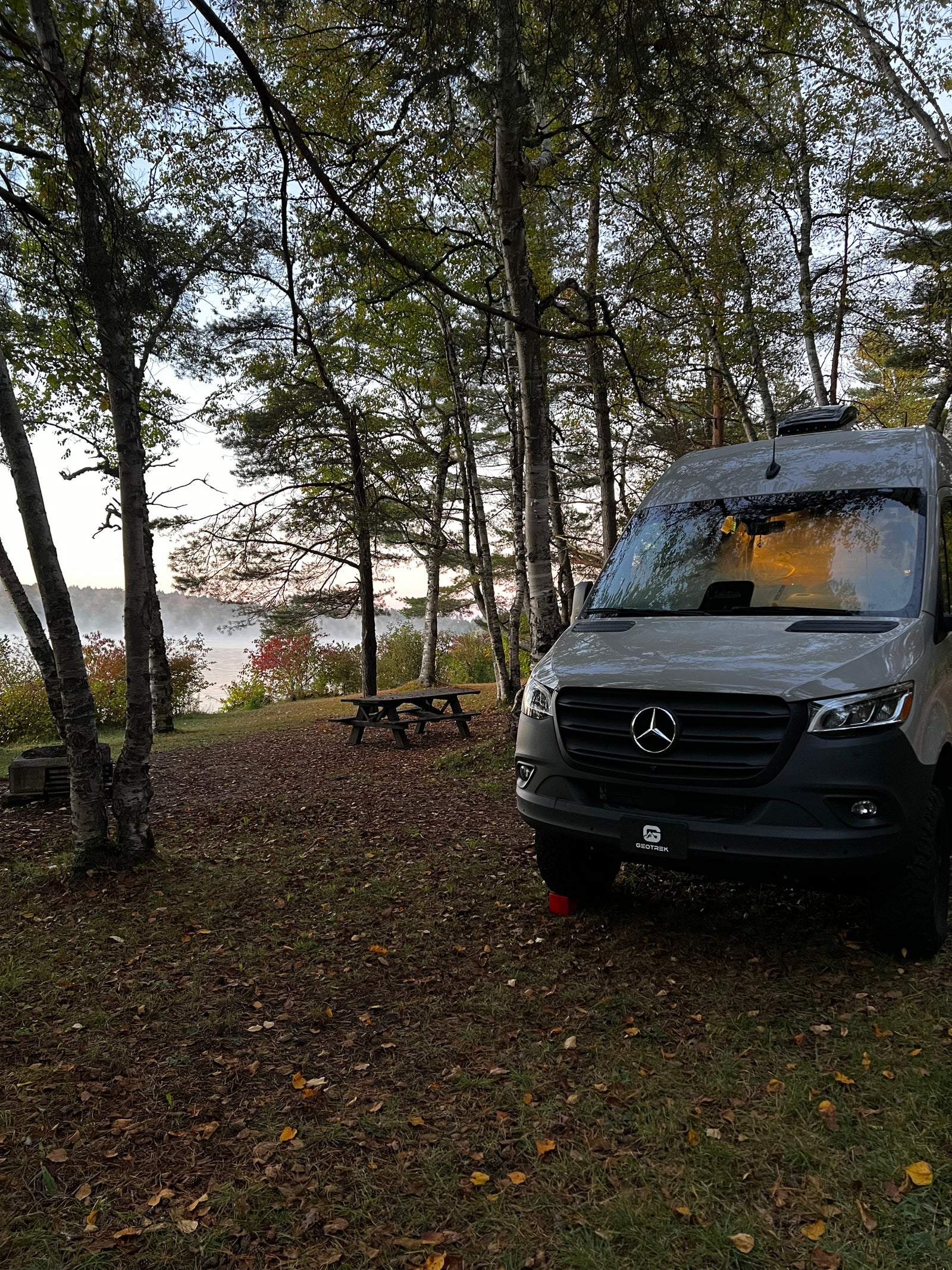 RV rental camper van parked by a misty lake, surrounded by autumn trees and a picnic table at a scenic campsite.