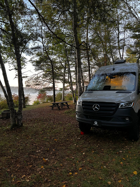 RV rental camper van parked by a misty lake, surrounded by autumn trees and a picnic table at a scenic campsite.