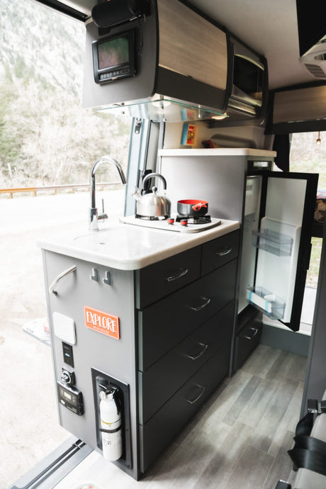 Inside an RV rental, showcasing a compact kitchen with sink, stove, fridge, and storage. Mountains visible outside.