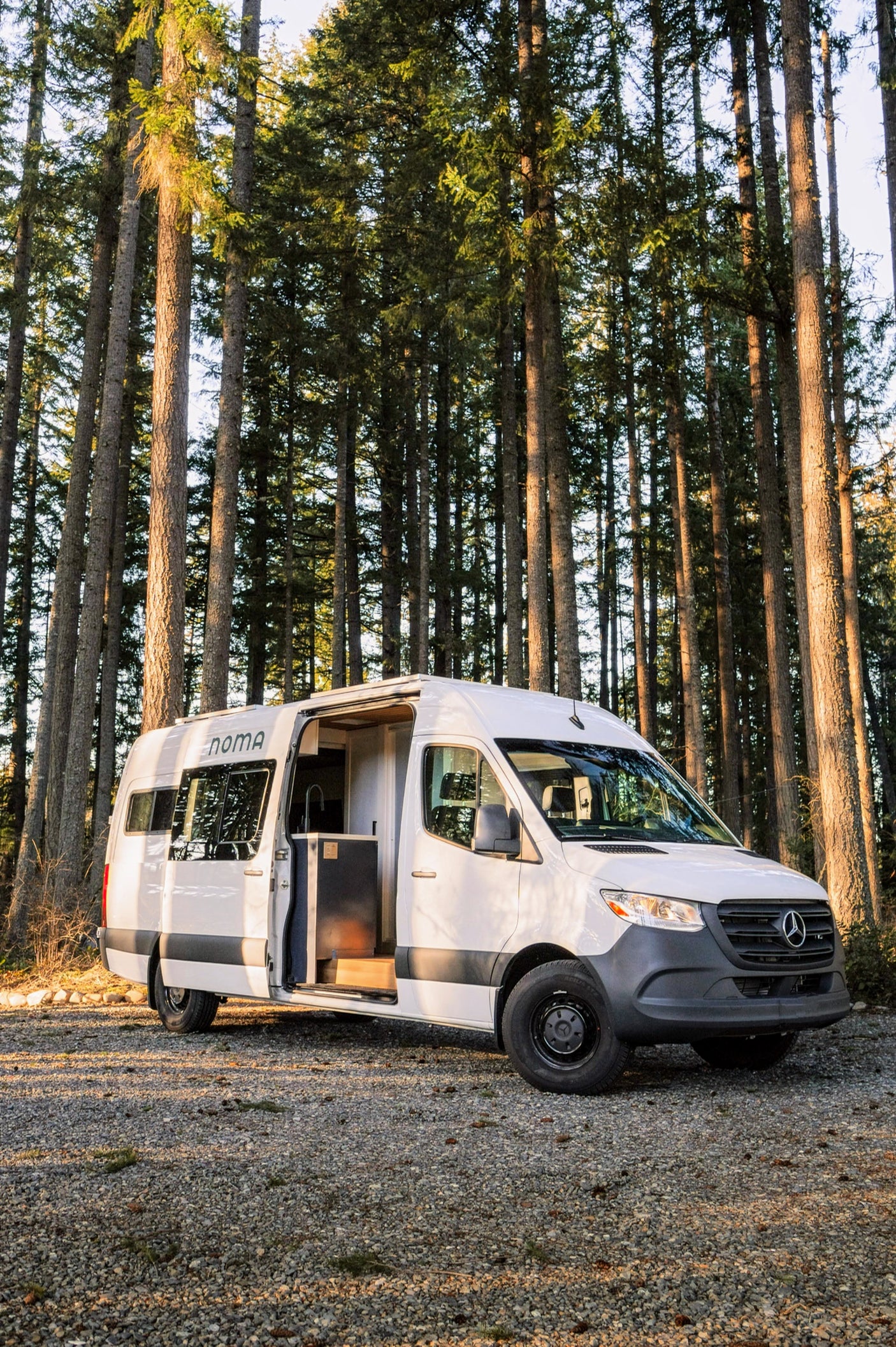 A modern NOMA rv rental camper van with its door open, parked on a gravel path amidst a dense forest.