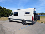 White camper van (RV rental) parked by a lake, with a ladder & spare tire on back. Perfect for travel adventures.