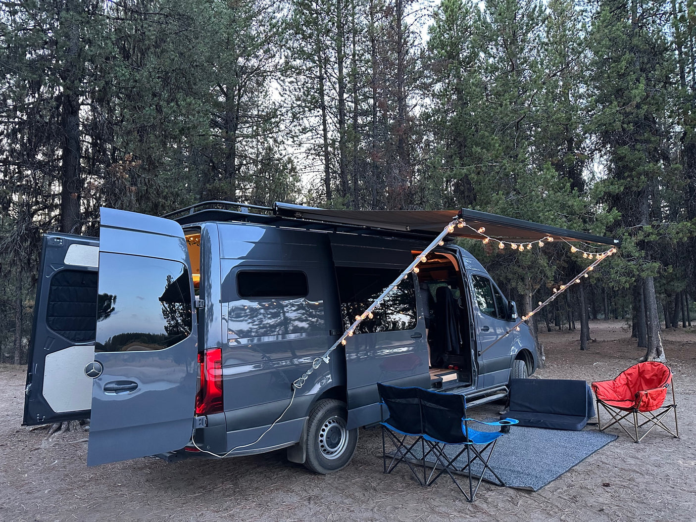 A dark grey camper van (RV rental) is parked in a forest, set up with an awning, string lights, and chairs for a cozy camp.