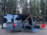 A dark grey camper van (RV rental) is parked in a forest, set up with an awning, string lights, and chairs for a cozy camp.