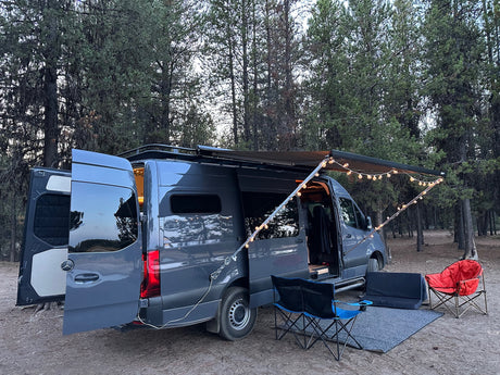 A dark grey camper van (RV rental) is parked in a forest, set up with an awning, string lights, and chairs for a cozy camp.